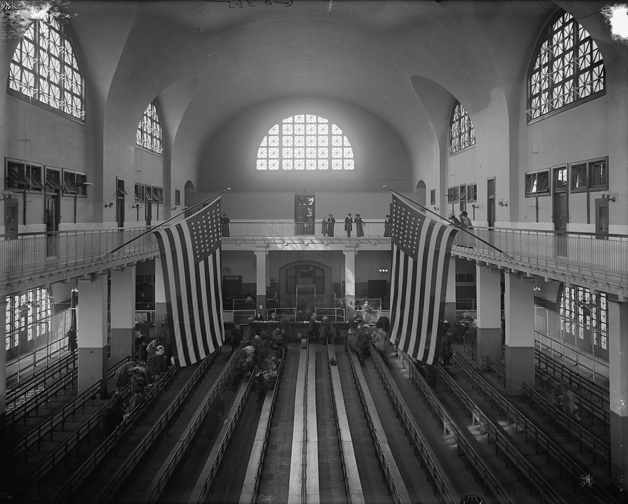 The Registry Room, or Great Hall, at Ellis Island, with rows of inspection benches, large American flags, and arched windows, photographed in the early 1900s