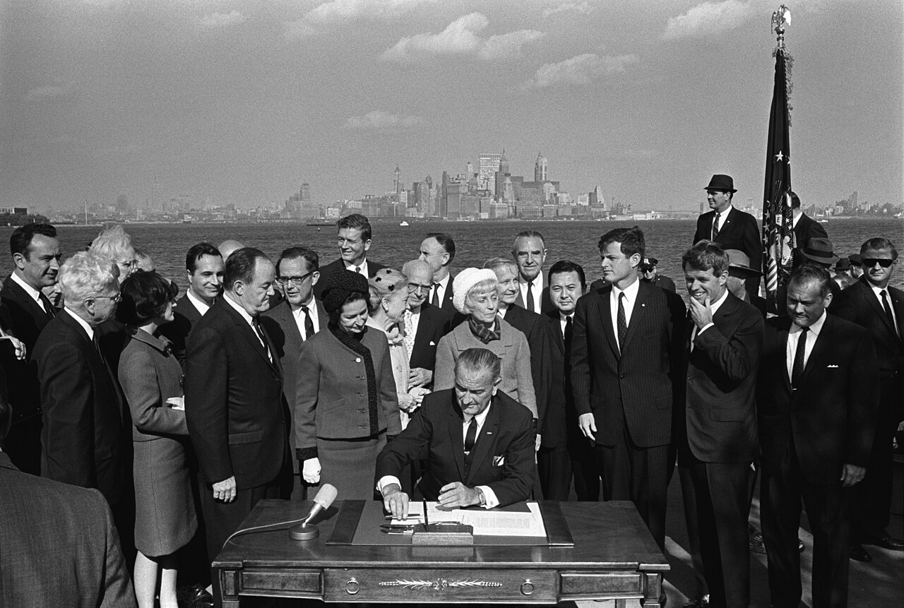 President Lyndon B. Johnson seated at a desk on Liberty Island signing the Immigration and Nationality Act of 1965, surrounded by Vice President Hubert Humphrey, Lady Bird Johnson, and members of Congress, with the Manhattan skyline visible across the harbor