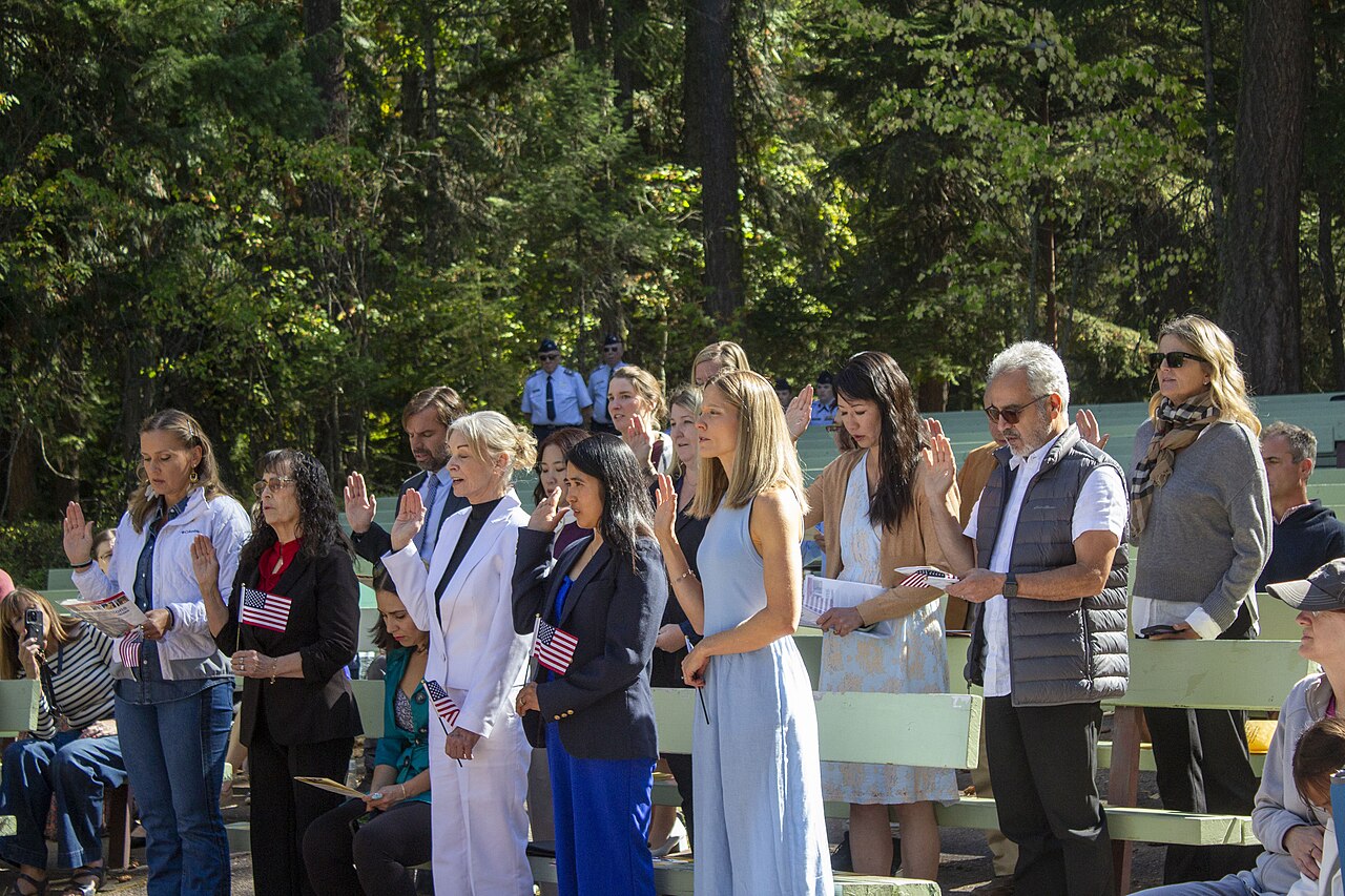 A row of new U.S. citizens with their right hands raised, taking the Oath of Allegiance at an outdoor naturalization ceremony